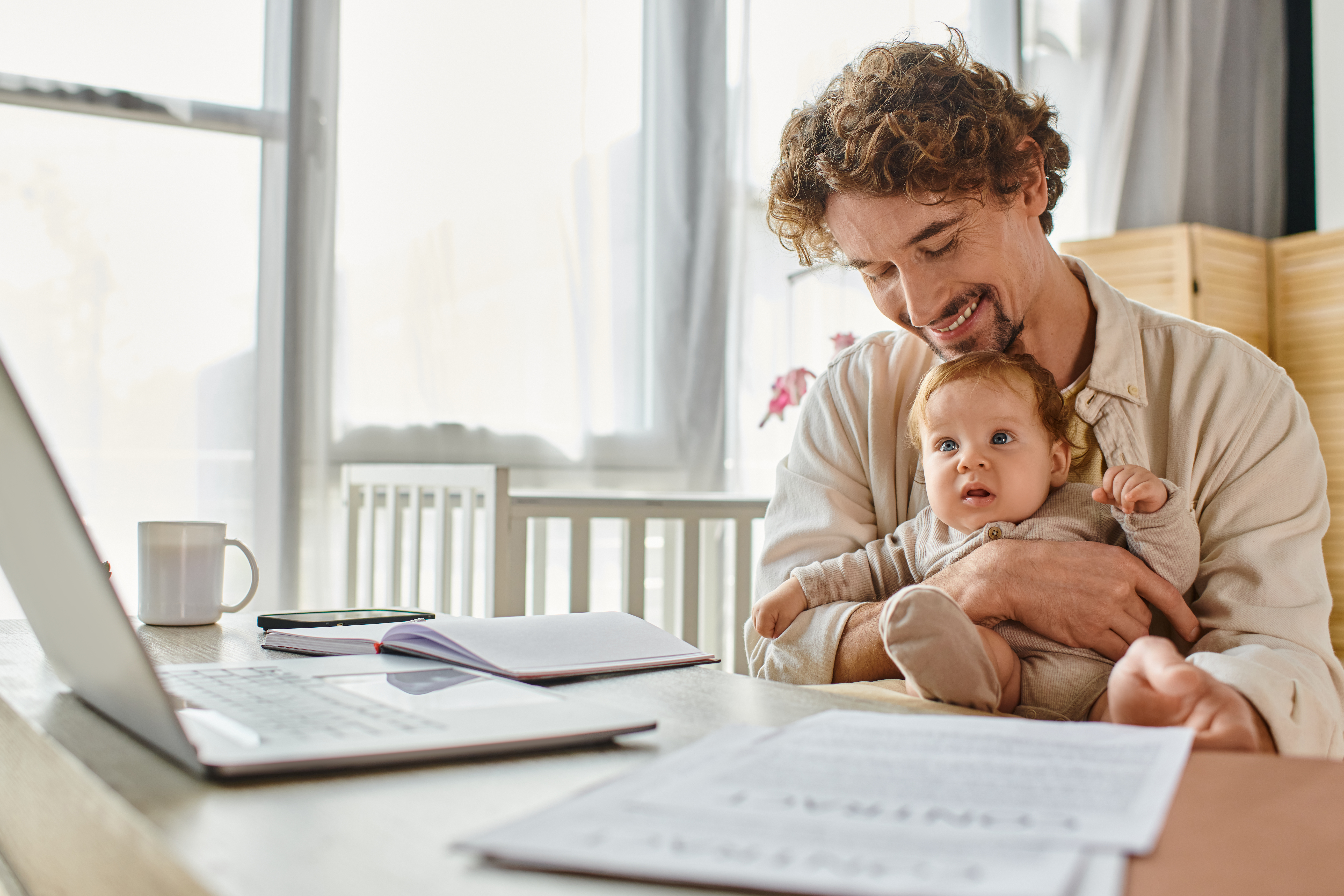 happy father holding infant son while working from home near papers and gadgets, work-life balance Illustrative photo